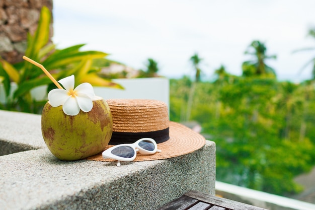 Cóctel de coco decorado plumeria, sombrero de paja y gafas de sol sobre la mesa.