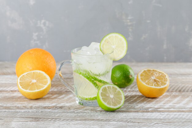 Cóctel de mojito helado en una taza con limas, naranja, vista lateral de limón en madera y pared de yeso