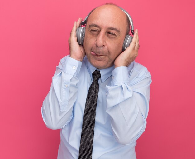 Codiciosos mirando al lado del hombre de mediana edad con camiseta blanca con corbata y auriculares aislados en la pared rosa