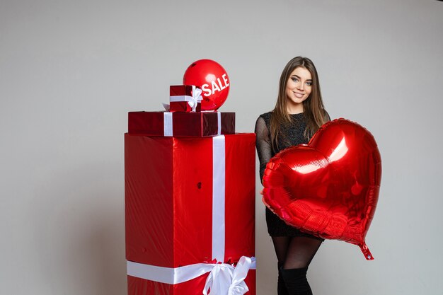 Colección de foto - morena, niña, en, vestido negro, tenencia, rojo, corazón, globo, posición, al lado de, envuelto, regalos Globo de aire con palabra de venta encima de regalos.