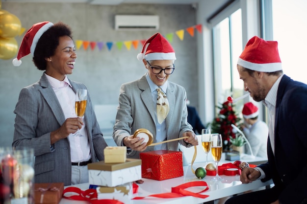 Colegas alegres divirtiéndose mientras envuelven regalos de Navidad en la oficina