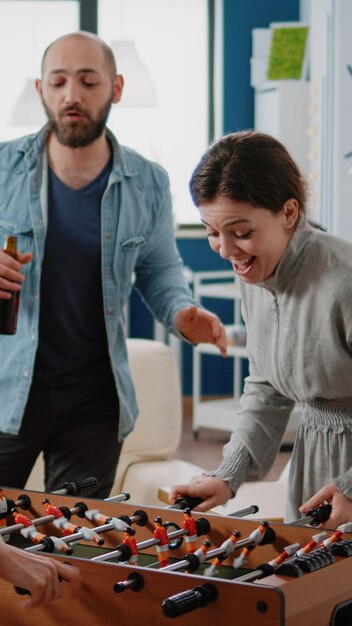 Colegas jugando futbolín y mujer ganando, divirtiéndose con futbolín después del trabajo. Compañeros de trabajo disfrutando de bebidas en la oficina, tomando botellas de cerveza y pizza para celebrar la fiesta