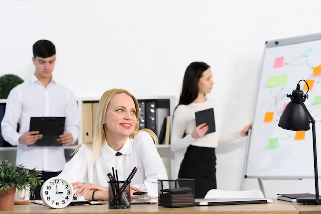 Colegas masculinos y femeninos que se colocan detrás de la empresaria sonriente que se sienta en el lugar de trabajo