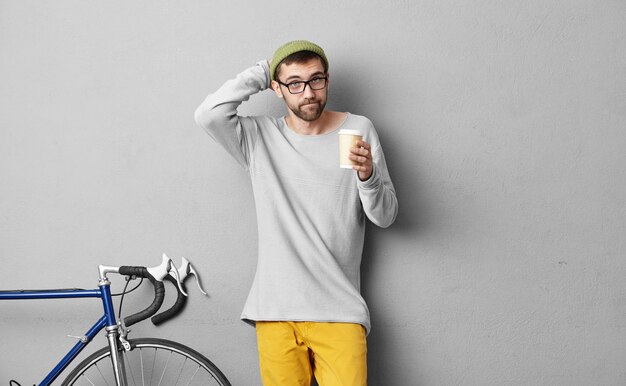 Colegial estirando la cabeza mientras mantiene café para llevar, tratando de recordar lo que debe llevar en su bolso, de pie cerca de la pared gris y la bicicleta. Hombre de picnic en bicicleta. Personas y descanso