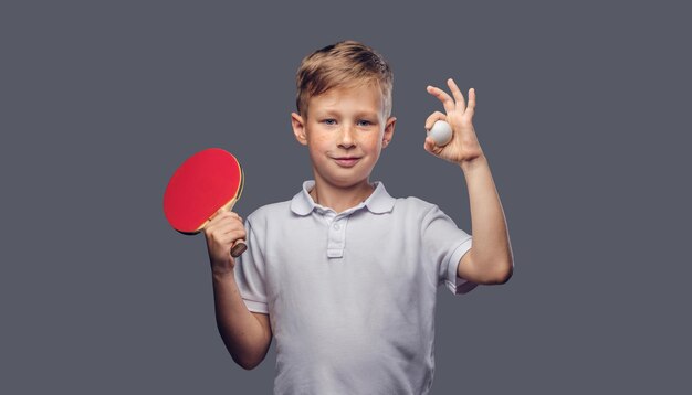 Un colegial pelirrojo vestido con una camiseta blanca sostiene una raqueta de ping-pong y una pelota en un estudio. Aislado sobre fondo gris.