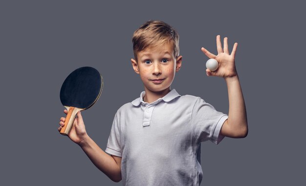 Un colegial pelirrojo vestido con una camiseta blanca sostiene una raqueta de ping-pong y una pelota en un estudio. Aislado sobre fondo gris.