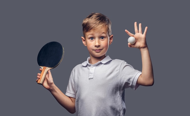 Un colegial pelirrojo vestido con una camiseta blanca sostiene una raqueta de ping-pong y una pelota en un estudio. Aislado sobre fondo gris.