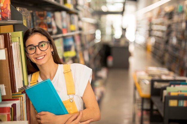 Colegiala adolescente feliz con libro cerca de estantes