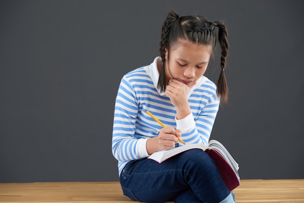 Colegiala asiática sentada en el escritorio, escribiendo en el libro de ejercicios con la barbilla apoyada en la mano