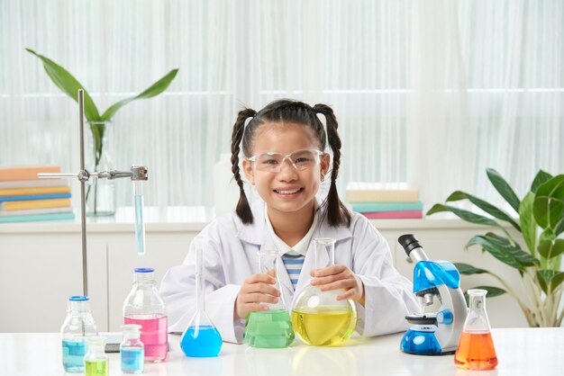 Colegiala asiática con trenzas sentado en el escritorio con microscopio y viales con líquidos coloridos