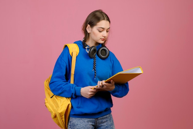 Colegiala con auriculares de pie sosteniendo carpeta amarilla leyendo