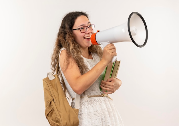 Colegiala bonita joven alegre con gafas y mochila sosteniendo libros hablando por altavoz aislado en blanco