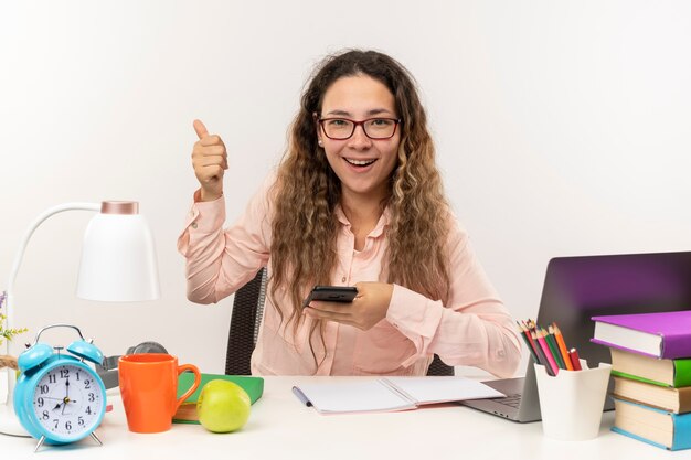 Colegiala bonita joven alegre con gafas sentado en el escritorio con herramientas escolares haciendo sus deberes sosteniendo el teléfono móvil y mostrando el pulgar hacia arriba aislado en blanco