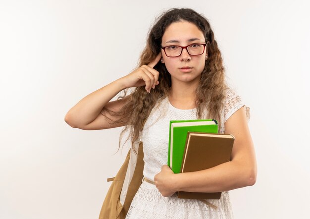 Colegiala bonita joven confiada con gafas y bolsa trasera sosteniendo libros poniendo el dedo en la oreja aislado en blanco