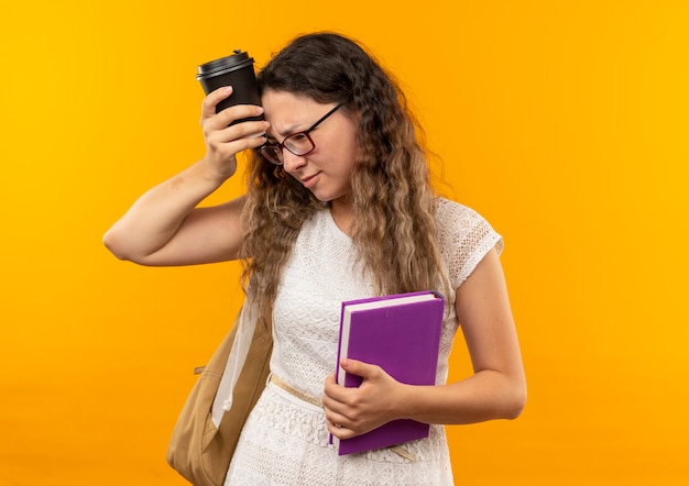 Colegiala bonita joven pensativa con gafas y bolsa trasera sosteniendo el libro mirando al lado tocando la cabeza con una taza de café de plástico aislada en la pared amarilla