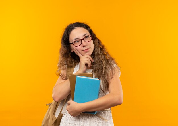 Colegiala bonita joven pensativa con gafas y bolsa trasera sosteniendo libros poniendo la mano en la barbilla mirando al lado aislado en la pared amarilla