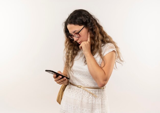 Colegiala bonita joven pensativa con gafas y bolso trasero usando su teléfono poniendo la mano en la barbilla aislada en la pared blanca