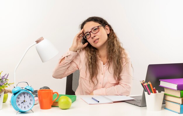 Colegiala bonita joven pensativa con gafas sentado en el escritorio con herramientas escolares haciendo sus deberes mirando al lado con la mano en la cabeza aislada en la pared blanca