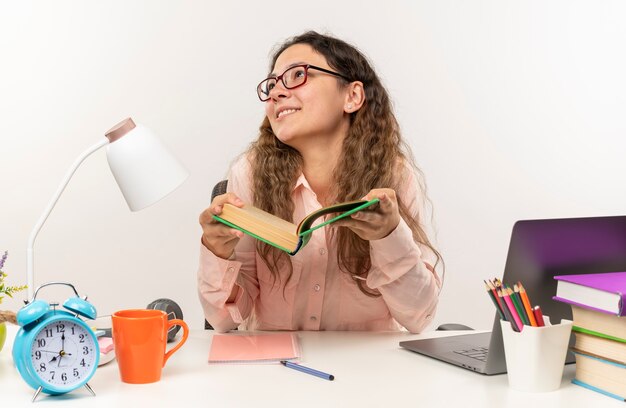 Colegiala bonita joven sonriente con gafas sentado en el escritorio con herramientas escolares haciendo sus deberes sosteniendo el libro mirando hacia arriba aislado en la pared blanca