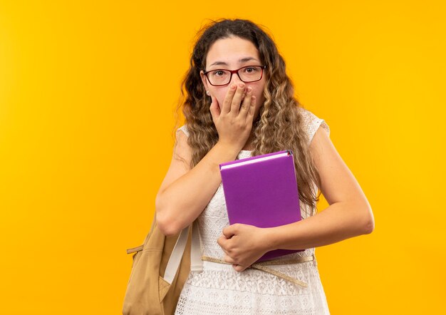 Colegiala bonita joven sorprendida con gafas y bolsa trasera sosteniendo el libro poniendo la mano en la boca aislada en la pared amarilla