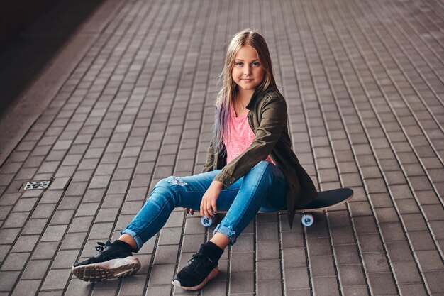 Colegiala con cabello rubio vestida con ropa de moda sentada en una patineta en un puente peatonal.