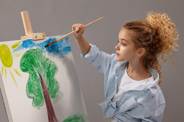 Una colegiala increíble con el pelo rubio rizado, vestida con una camisa azul y una camiseta blanca, está pintando con un pincel de acuarela en un caballete, de pie sobre un fondo gris.