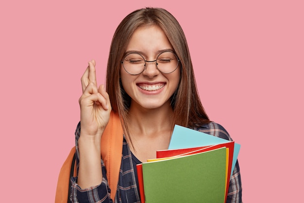 Colegiala intensa y alegre con amplia sonrisa, cruza los dedos para la buena suerte, tiene expresión satisfecha, mantiene los ojos cerrados, lleva mochila,