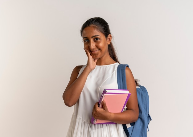 Colegiala joven complacida con bolsa trasera sosteniendo el cuaderno y poniendo la mano en la mejilla en blanco