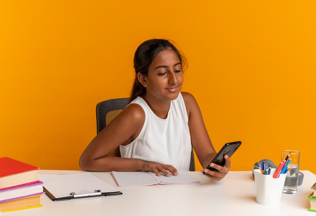 Colegiala joven complacida sentada en un escritorio con herramientas escolares sosteniendo y mirando el teléfono aislado en la pared naranja