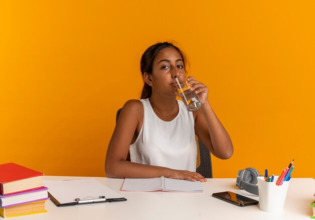 Colegiala joven sentada frente al escritorio con herramientas escolares agua potable