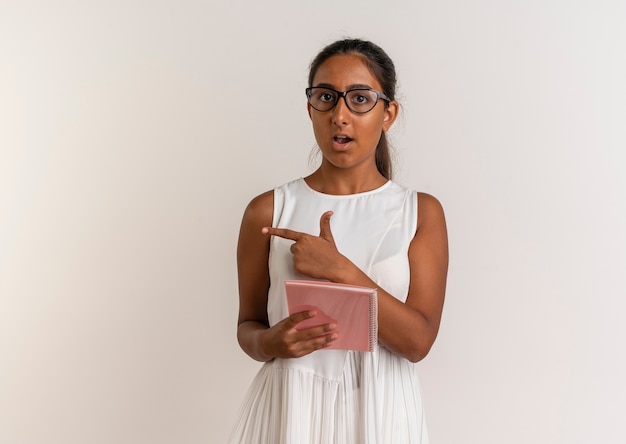 Foto gratuita colegiala joven sorprendida con gafas sosteniendo el cuaderno y apunta al lado aislado en la pared blanca con espacio de copia