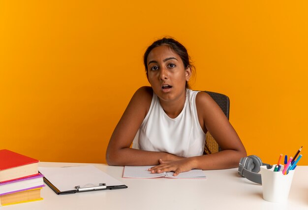 Colegiala joven sorprendida sentada en el escritorio con herramientas escolares en naranja