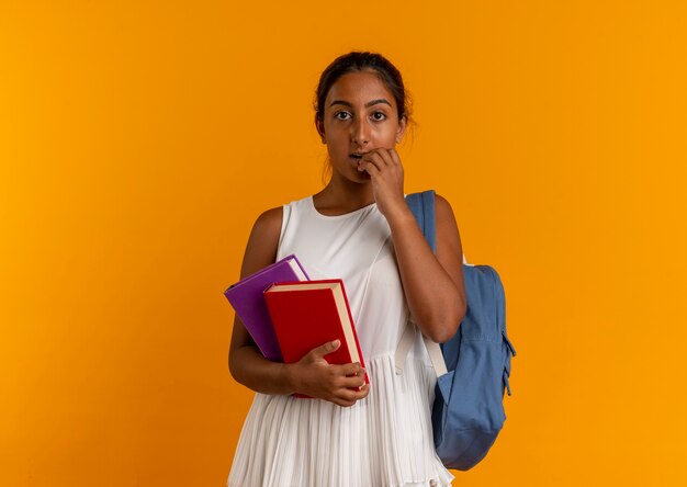 Colegiala joven sorprendida vistiendo bolsa trasera sosteniendo libros y poniendo la mano en la boca