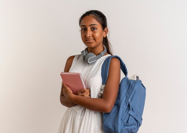 Colegiala joven vistiendo bolsa trasera y auriculares con portátil en blanco