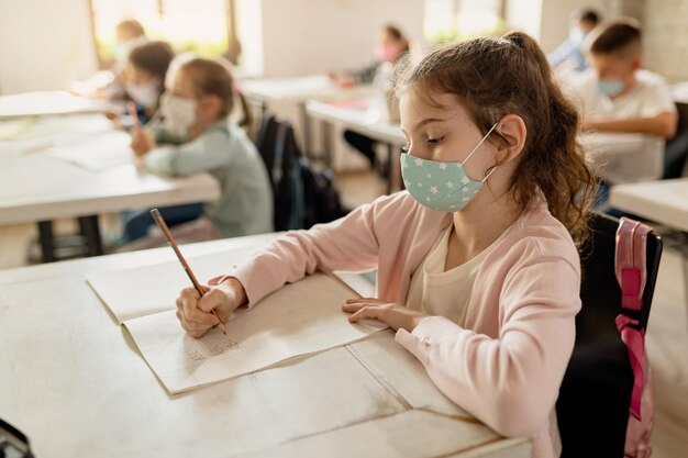 Colegiala con mascarilla protectora escribiendo en un cuaderno durante una conferencia en el aula