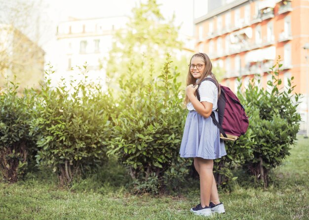 Colegiala con mochila de pie delante de arbustos sonriendo