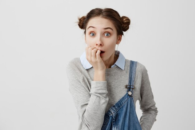 Colegiala con el pelo en doble moño mirando asustada mordiéndose las uñas en tensión. Emociones negativas de una mujer con ojos que salen en problemas mientras dejaba su plancha tapada. Reacciones humanas