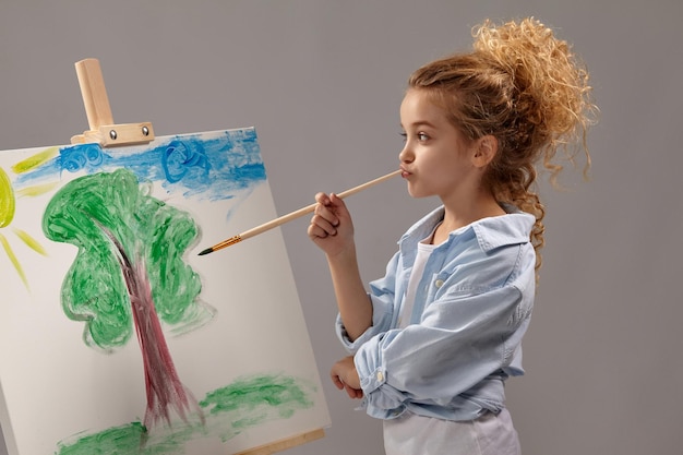 Una colegiala pensativa con el pelo rubio, vestida con una camisa azul y una camiseta blanca, está pintando con un pincel de acuarela en un caballete, de pie sobre un fondo gris.