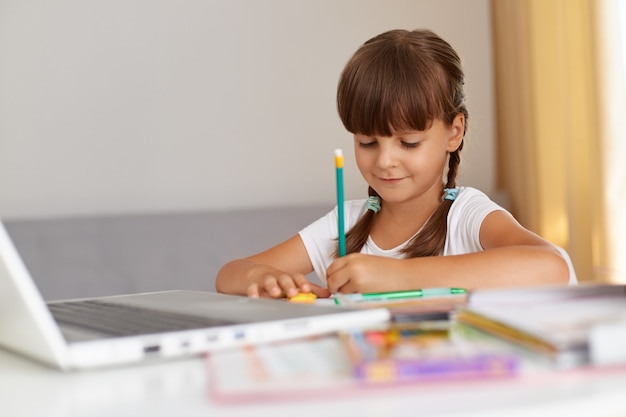 Colegiala positiva y guapa con ropa informal, escribiendo en un cuaderno, con un estado de ánimo positivo, sentada a la mesa en la sala de estar, educación en línea.