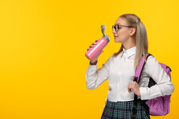 Colegiala rubia del día mundial del libro en lindo uniforme bebiendo de termo rosa