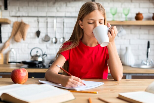 Colegiala sosteniendo la taza haciendo la tarea en la mesa de la cocina