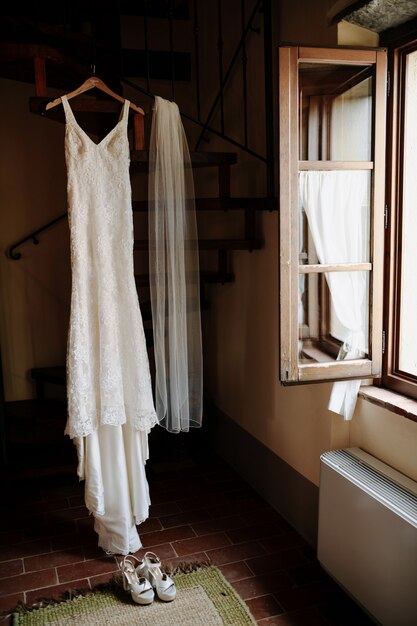Colgar el elegante vestido de novia y el velo de novia en la habitación cerca de la ventana abierta