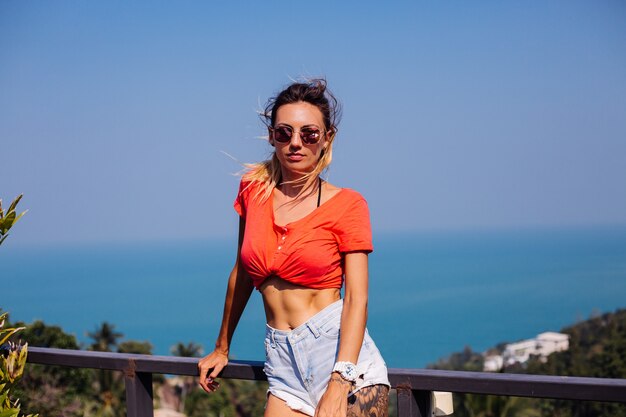 Colocar bronceada tatuada mujer europea caucásica con cuerpo deportivo y abs, en pantalones cortos de jean y camiseta roja naranja