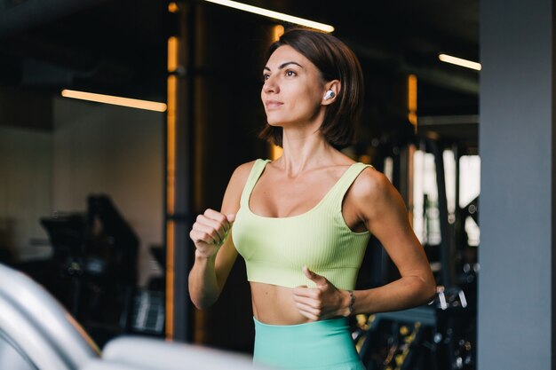 Colocar hermosa mujer caucásica en ropa deportiva adecuada en el gimnasio corriendo trotar en cinta con auriculares inalámbricos en los oídos