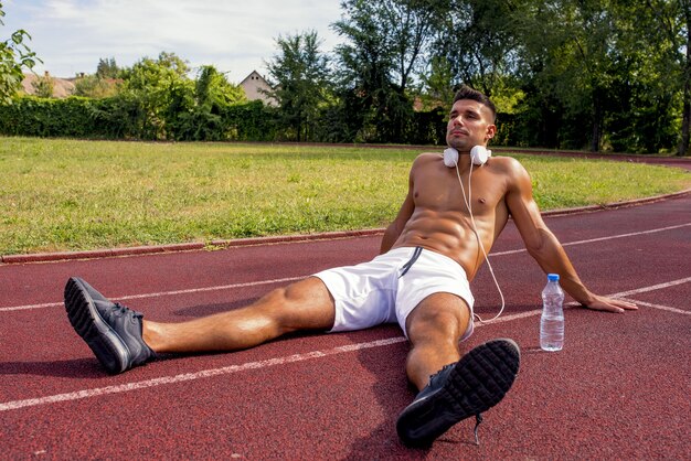Colocar hombre sin camisa con auriculares sentado en un descanso de hacer ejercicio en una pista de atletismo