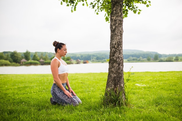 Colocar joven mujer haciendo yoga en el parque cerca del lago y el árbol
