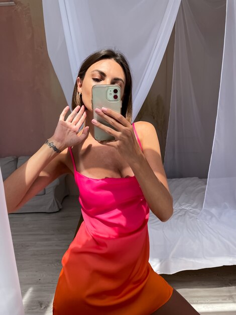 Colocar mujer bronceada en lindo vestido de seda naranja de verano brillante en casa toma foto selfie en teléfono