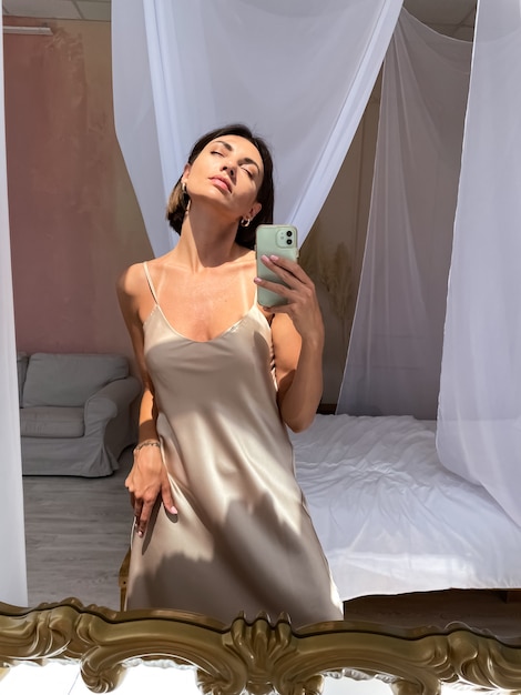 Colocar mujer bronceada en romántico vestido de seda beige en casa toma foto selfie en teléfono en el espejo