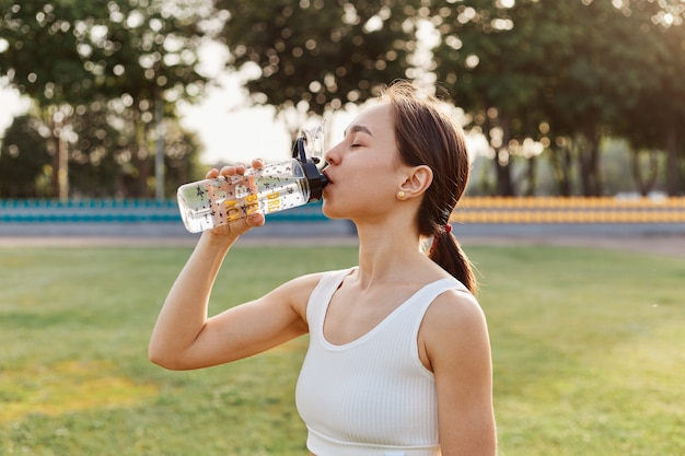 Colocar a una mujer de cabello oscuro bebiendo agua de su botella de agua en el estadio justo antes de comenzar a entrenar, hacer ejercicio al aire libre en verano, estilo de vida saludable.