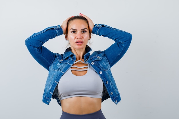 Foto gratuita colocar a la mujer con las manos en la cabeza en la parte superior de la cosecha, chaqueta de jean, leggings y aspecto serio. vista frontal.